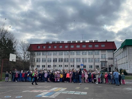 Stojące dzieci na boisku szkolnym z klas 1-4 wraz z Paniami. W tle budynek szkoły podstawowej nr 5.
