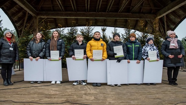 Na zdjęciu znajdują się dzieci wraz z nauczycielkami. Wszyscy stoją ubrani w kurtkach, w tle są choinki. Dzieci trzymają w rękach dyplomy oraz nagrody otrzymane za udział w konkursie.