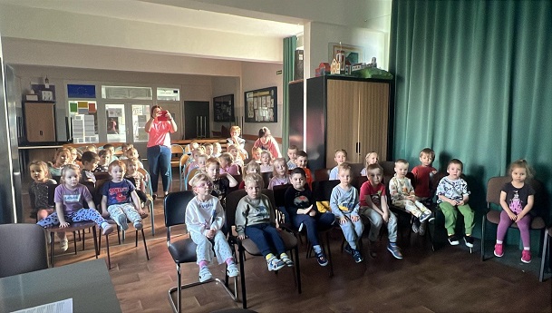 Na zdjęciu znajdują się przedszkolaki. Dzieci siedzą na krzesełkach w czytelni i uważnie oglądają przedstawienie.