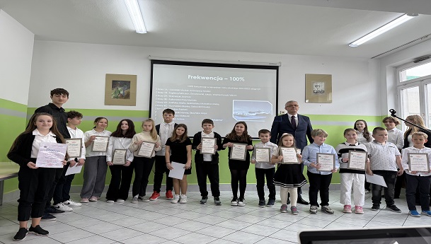 yrektor szkoły stoi wraz z uczniami klas 1-8, którzy trzymają dyplomy za 100% frekwencję.