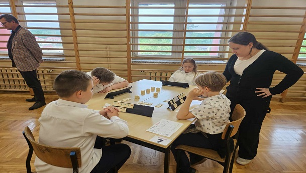 Uczniowie siedzący przy stoliku grający w Rummikub, Wicedyrektor przy stoliku oglądający uczniów podczas gry